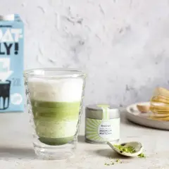 Warstwowy napój z zielonej herbaty matcha w szklance, obok puszka z napisem "PREMIUM MATCHA" i bambusowa miotełka.