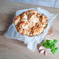 Pyszny chlebek rwany z ziołami, ułożony w okrągłej formie, obok świeża bazylia i czosnek.