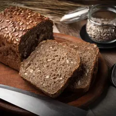 Chleb proteinowy z pestkami słonecznika na drewnianej desce, obok nasion w słoiku i łopatce.