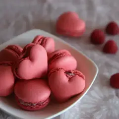 Różowe makaroniki francuskie w kształcie serc ułożone na talerzyku, obok kilka malin.
