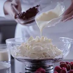 Płynna biała czekolada spływa na pokruszoną białą czekoladę w misce, tworząc bazę do ganache z białej czekolady.