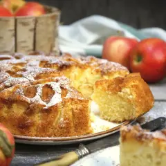 Pyszne, domowe ciasto z jabłkami, posypane cukrem pudrem, prezentuje się apetycznie obok świeżych jabłek.