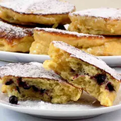 Puszyste racuchy na kefirze jak u babci, posypane cukrem pudrem, z jagodami w środku. Smak dzieciństwa na talerzu.