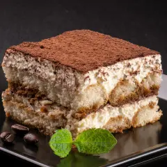 Idealne, najprostsze tiramisu z kremem i biszkoptami, posypane kakao. Obok leżą ziarna kawy i listek mięty.