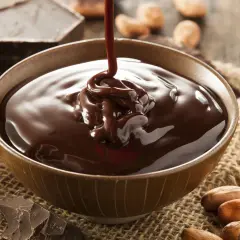 Gęsta, prosta polewa czekoladowa z czekolady płynie do miseczki. Wokół kawałki czekolady i ziarna kakaowca.