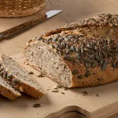 Chleb orkiszowy z pestkami dyni i sezamu, pokrojony na desce, obok nóż i koszyk.