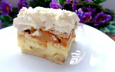 Szybkie ciasto z bananami i budyniem, przełożone kremem i bitą śmietaną, prezentuje się apetycznie na talerzu, w otoczeniu fioletowych kwiatów.
