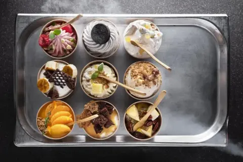 Dziewięć deserów typu parfait na tacy: malinowy, oreo, z orzechami, bananowy, pomarańczowy i inne.