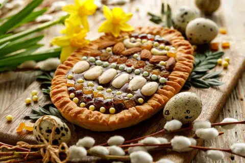 Wielkanocny mazurek ozdobiony bakaliami, orzechami i migdałami, otoczony pisankami i baziami.