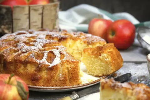 Pyszne, domowe ciasto z jabłkami, posypane cukrem pudrem, prezentuje się apetycznie obok świeżych jabłek.