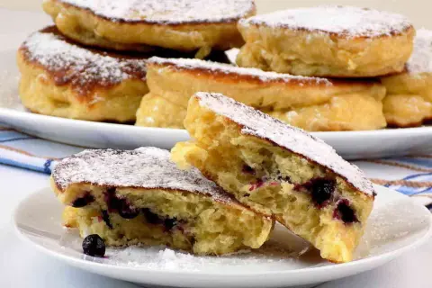 Puszyste racuchy na kefirze jak u babci, posypane cukrem pudrem, z jagodami w środku. Smak dzieciństwa na talerzu.