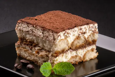 Idealne, najprostsze tiramisu z kremem i biszkoptami, posypane kakao. Obok leżą ziarna kawy i listek mięty.