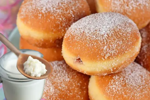 Szybkie pączki z piekarnika, oprószone cukrem pudrem, obok słoiczka z jogurtem i łyżki z jego zawartością.