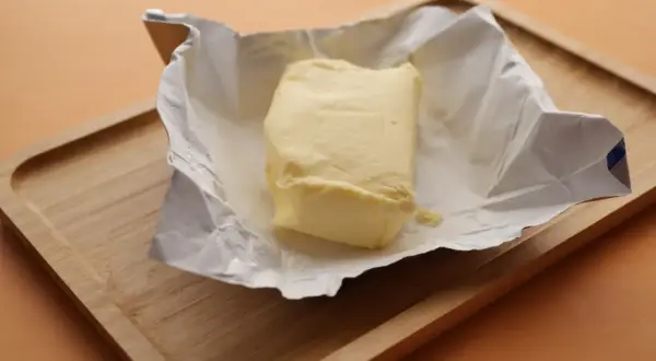 Kostka masła w papierku na drewnianej desce. Czy masło można mrozić? Tak, można je zamrozić, by zachować świeżość.