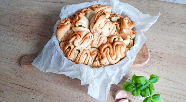 Pyszny chlebek rwany z ziołami, ułożony w okrągłej formie, obok świeża bazylia i czosnek.