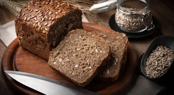 Chleb proteinowy z pestkami słonecznika na drewnianej desce, obok nasion w słoiku i łopatce.