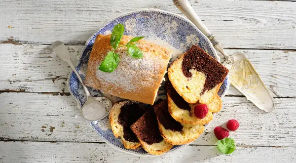 Babka piaskowa przepis babci: pyszne ciasto marmurkowe, posypane cukrem pudrem, z malinami i miętą na talerzu.