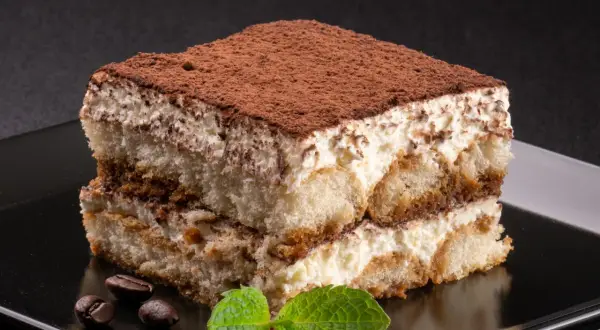 Idealne, najprostsze tiramisu z kremem i biszkoptami, posypane kakao. Obok leżą ziarna kawy i listek mięty.