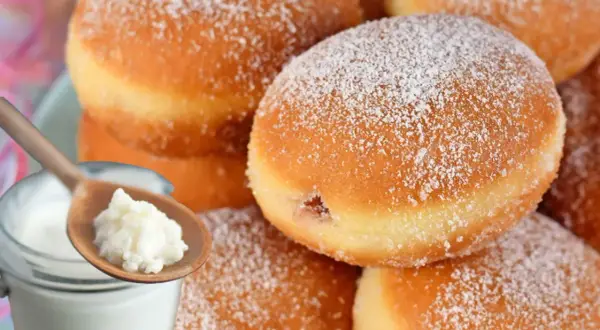 Szybkie pączki z piekarnika, oprószone cukrem pudrem, obok słoiczka z jogurtem i łyżki z jego zawartością.