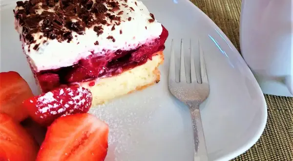 Pyszne ciasto z galaretką i truskawkami, udekorowane bitą śmietaną i czekoladą. Obok świeże truskawki i widelec.
