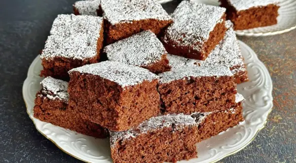 Szybkie i proste ciasto czekoladowe pokrojone w kostkę, posypane cukrem pudrem, ułożone na białym talerzu.