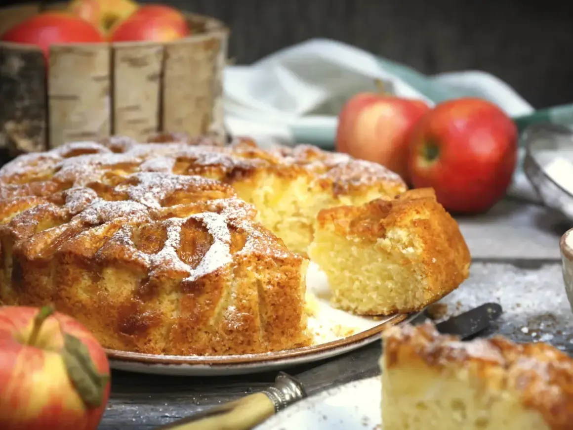Pyszne, domowe ciasto z jabłkami, posypane cukrem pudrem, prezentuje się apetycznie obok świeżych jabłek.