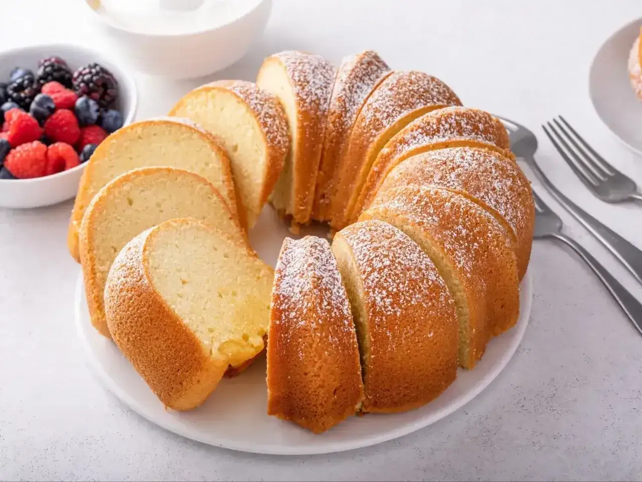 Puszysta babka z cukrem pudrem, pokrojona w plastry, obok miseczki z owocami leśnymi i śmietaną.