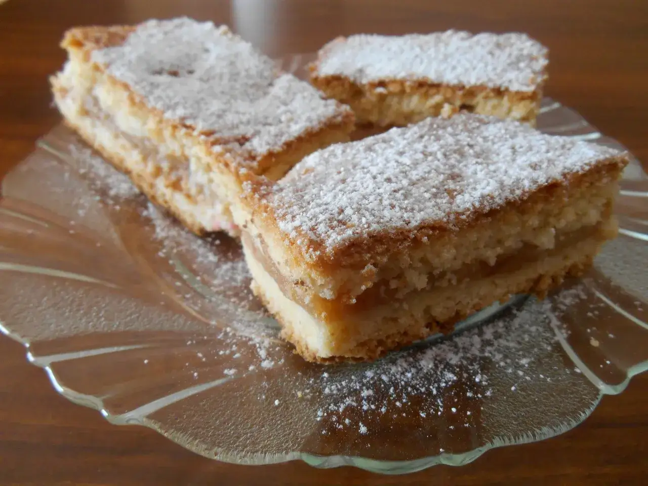 Trzy kawałki najprostszego jabłecznika, posypane cukrem pudrem, na szklanym talerzu.