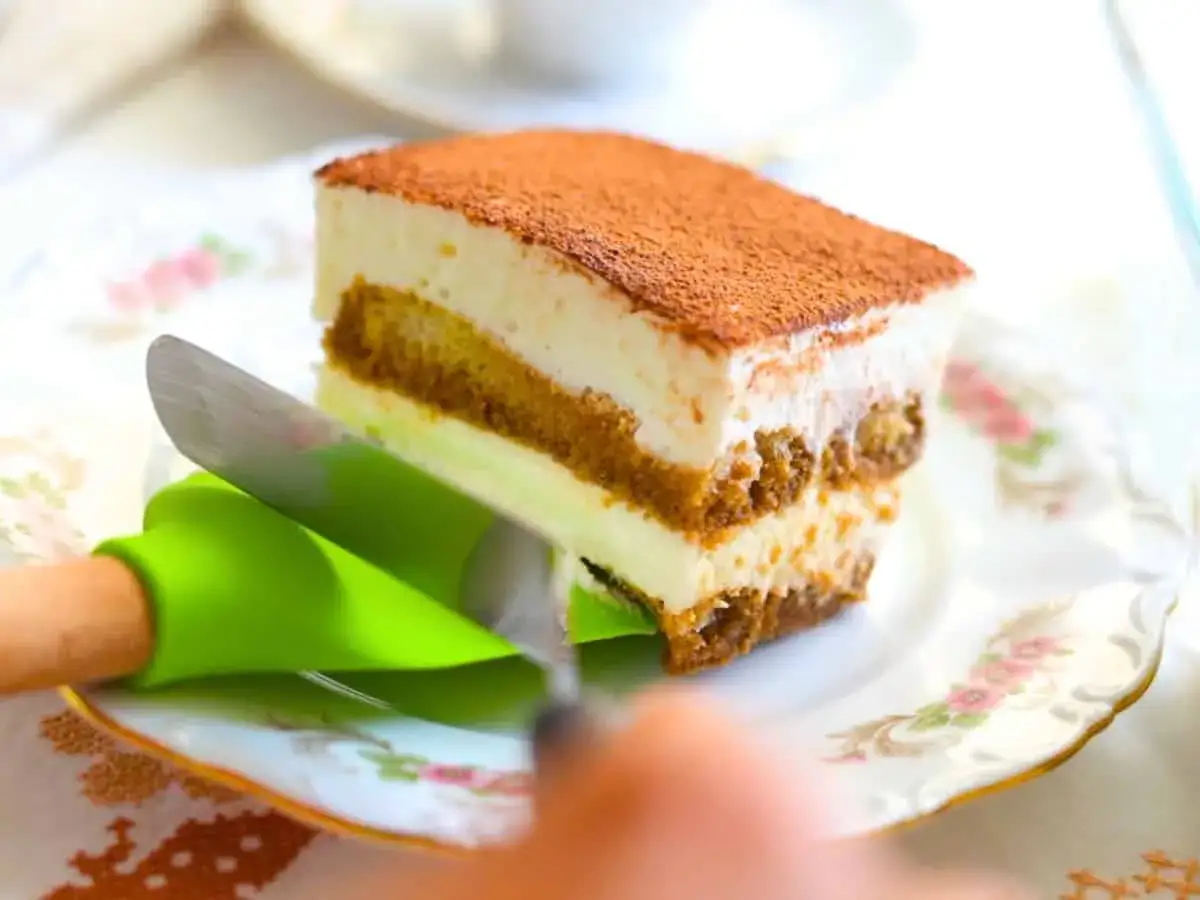 Kawałek najprostszego tiramisu, z kremową warstwą i biszkoptami, posypany kakao. Krojenie ułatwia zielona łopatka.
