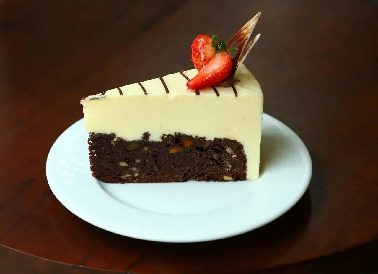 Kawałek ciasta brownie z orzechami, pokryty kremowym ganache z białej czekolady, ozdobiony truskawką i czekoladowymi wiórkami.