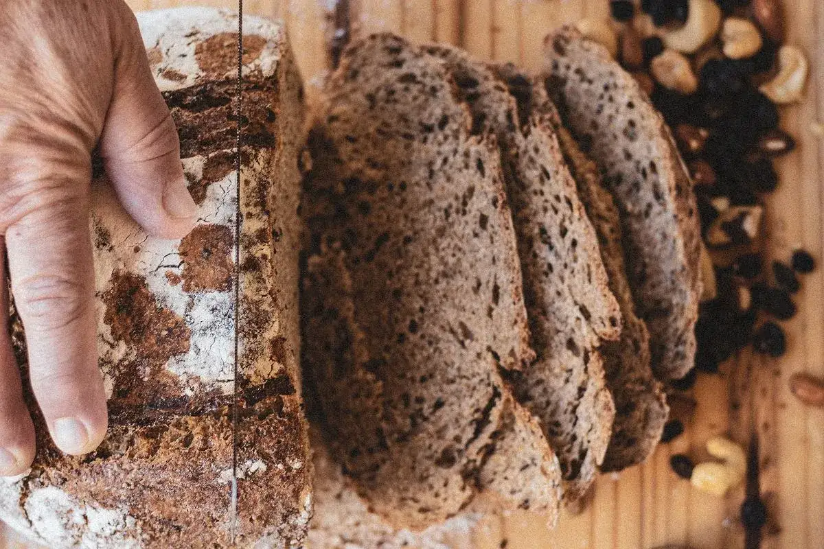 Ręka kroi bochenek zakwasu żytniego. Kromki chleba leżą obok, a w tle garść bakalii.