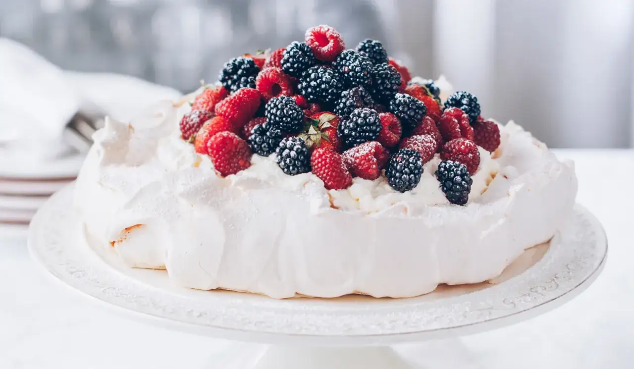 Beza pavlova która zawsze wychodzi, udekorowana bitą śmietaną i mnóstwem świeżych malin i jeżyn. Idealny deser na każdą okazję.