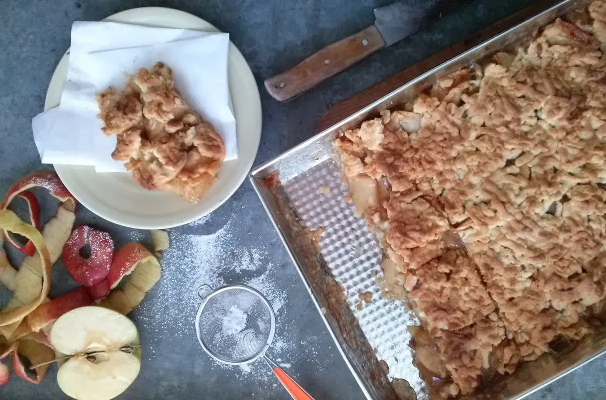 Pyszna szarlotka przepis na dużą blachę, świeżo upieczona, z jabłkami i kruszonką. Obok jabłka, skórki i sitko z cukrem pudrem.