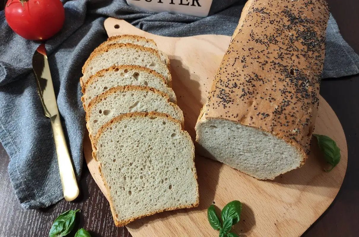 Chleb żytni na drożdżach, pokrojony w kromki, z makiem na skórce. Obok pomidor, nóż i listki bazylii.