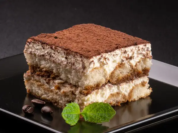 Idealne, najprostsze tiramisu z kremem i biszkoptami, posypane kakao. Obok leżą ziarna kawy i listek mięty.