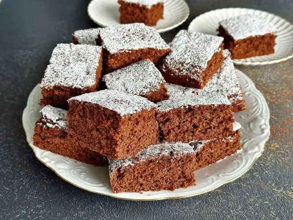 Szybkie i proste ciasto czekoladowe pokrojone w kostkę, posypane cukrem pudrem, ułożone na białym talerzu.